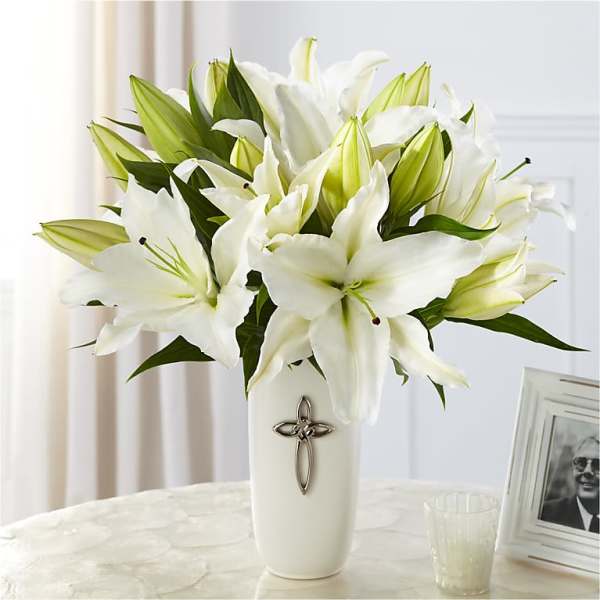 White lilies arranged in a tall white vase with a cross emblem