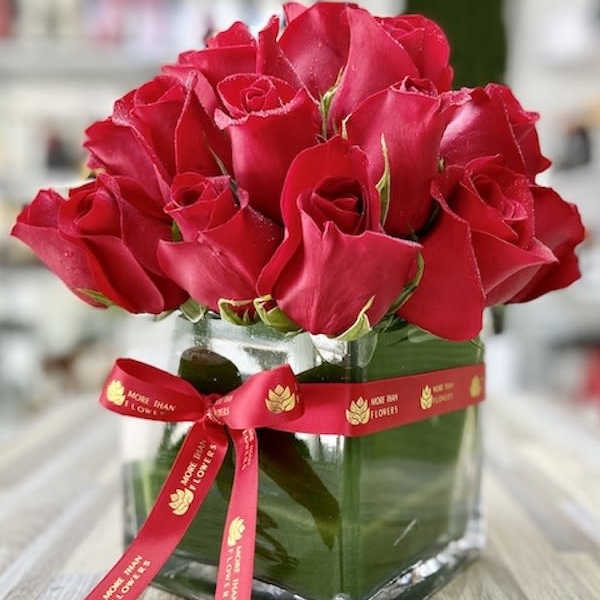 Compact arrangement of red roses in a clear glass cube vase with a red printed ribbon