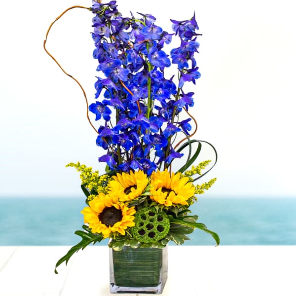 Tall blue delphinium and yellow sunflowers arranged in a clear rectangular glass vase