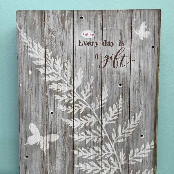 Rustic gray wooden sign with white foliage, butterflies, and the words "Every day is a gift".