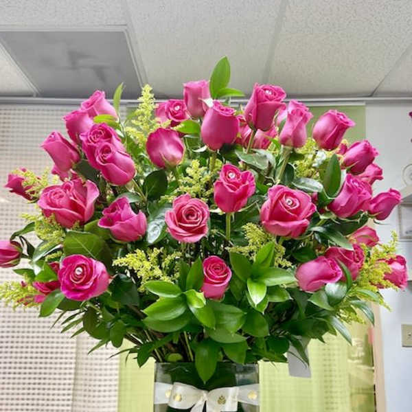 Tall arrangement of bright pink roses with filler flowers in a square glass vase with a white ribbon