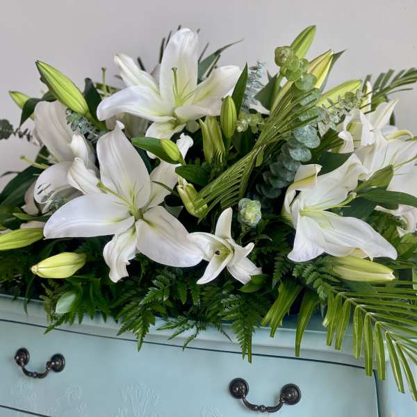 Low arrangement of white lilies with buds and abundant foliage on a pale blue dresser