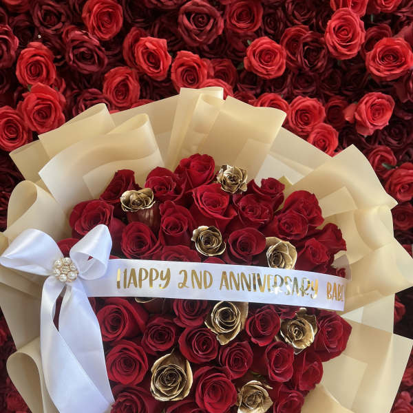 Round bouquet of red and gold roses wrapped in cream paper with a white bow and printed anniversary ribbon