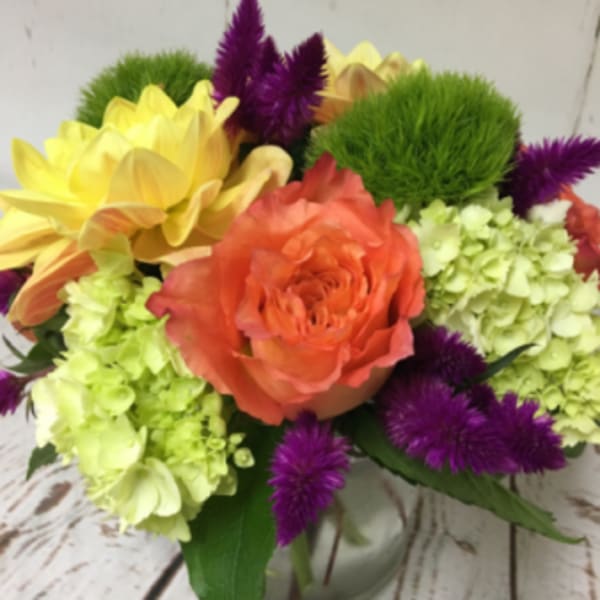 Low arrangement of orange rose, green hydrangea, yellow blooms and purple flowers in a clear glass vase