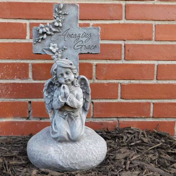 Gray angel statue kneeling before a cross with flowers and the words Amazing Grace