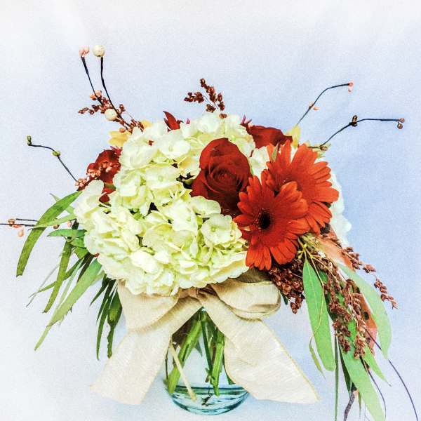 Low glass vase arrangement with white hydrangeas, red roses, and orange gerbera daisies tied with a cream bow.