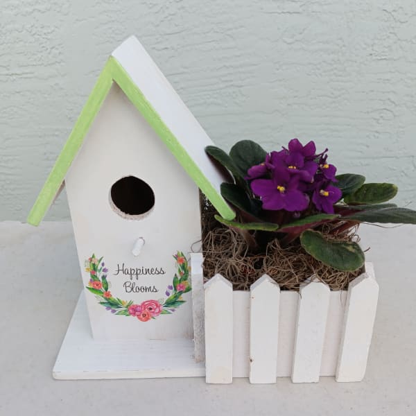 White birdhouse planter with purple flowers in a small picket fence box