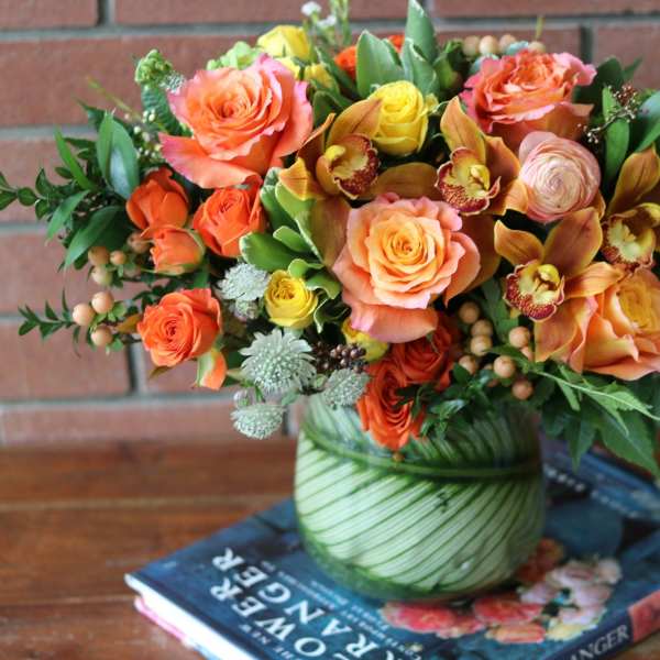 Low arrangement of orange and yellow roses with orchids in a round glass vase wrapped with green leaves