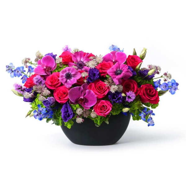 Low arrangement of hot pink roses, purple orchids, and blue flowers in a black bowl vase