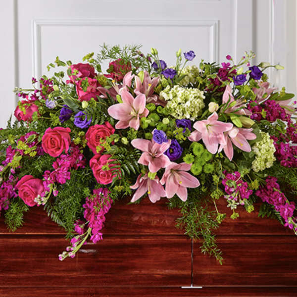 Bright pink lilies, roses, and purple blooms arranged as a casket spray on a wooden casket