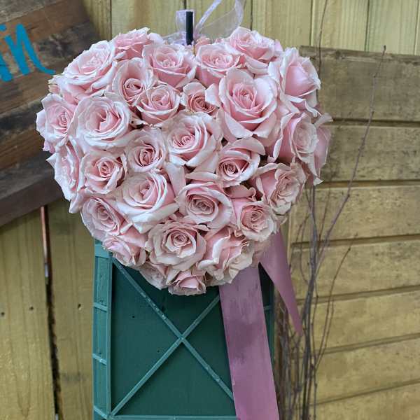 Heart-shaped arrangement of light pink roses with a pink ribbon on a green easel stand.