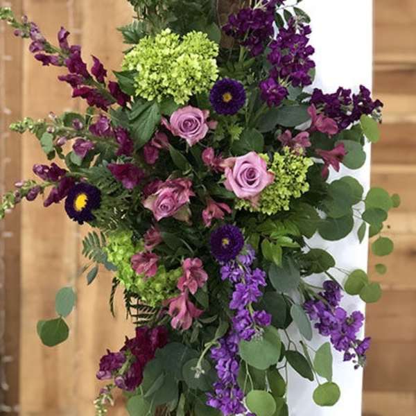 Asymmetrical purple and pink flower spray with green hydrangeas cascading from a wooden post.