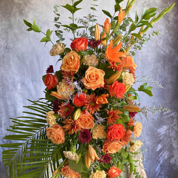 Tall orange and peach standing floral spray with roses and lilies on an easel.