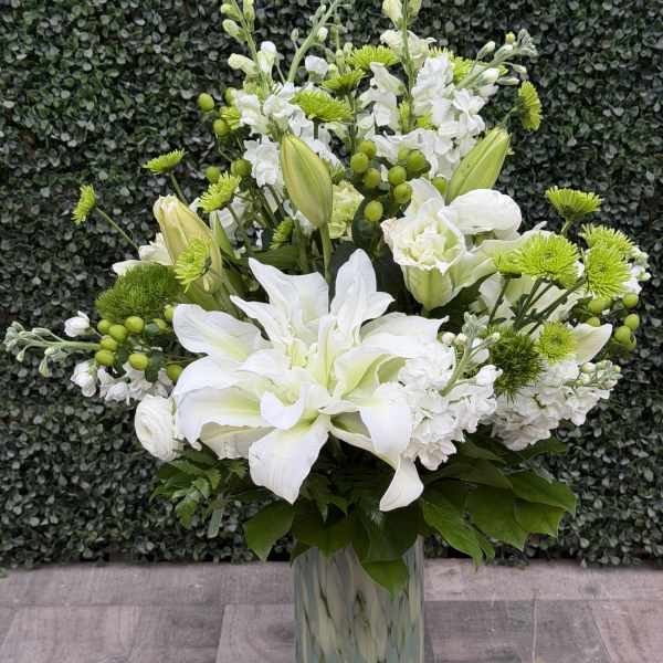 Tall white and green floral arrangement with lilies and mums in a patterned glass vase