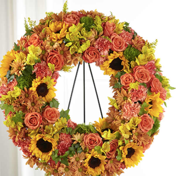 Standing floral wreath of yellow sunflowers, orange roses, and mixed warm-toned blooms on a metal easel