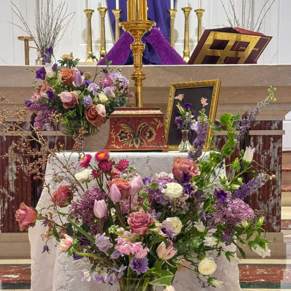 Large purple and pink mixed flower arrangements on a church altar with candles and framed photo