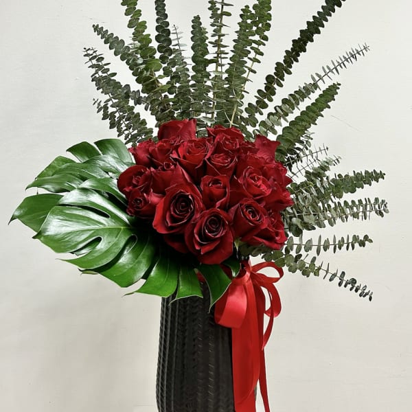 Tall black vase with red roses, large tropical leaf, and radiating eucalyptus stems.