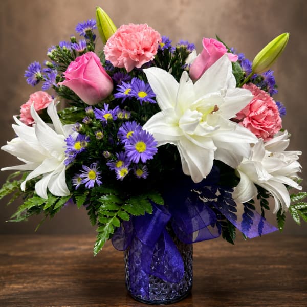 Pink roses, white lilies, and purple daisies in a glass vase with a purple ribbon