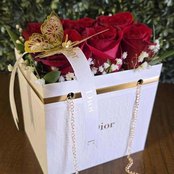 Red roses arranged in a white Dior gift box with a ribbon and gold butterfly decoration.