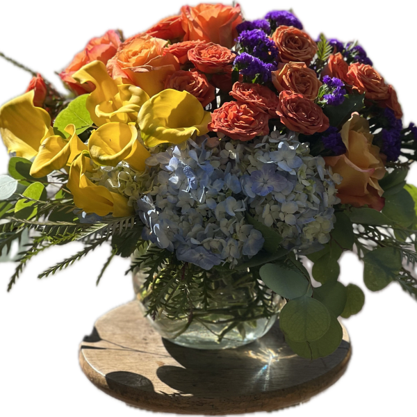 Colorful bouquet with yellow calla lilies, orange roses, blue hydrangea, and purple accents in a glass vase