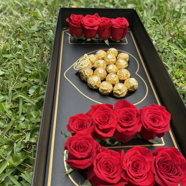 Black gift box with red roses and gold-wrapped chocolates on grass