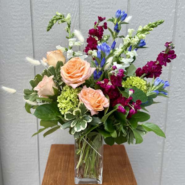 Mixed bouquet with peach roses, blue blooms, and magenta flowers in a glass vase