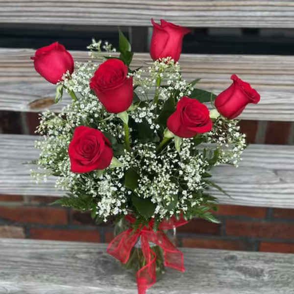 Glass vase of six red roses with white filler flowers and a red bow on a wooden bench