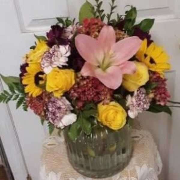 Mixed bouquet with a pink lily, yellow roses, and purple flowers in a glass vase