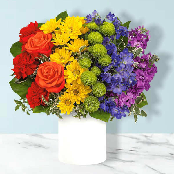 Rainbow bouquet in a white vase with roses, daisies, and purple blooms