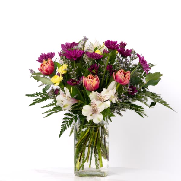 Mixed bouquet of purple daisies, pink tulips, and white blooms in a clear glass vase