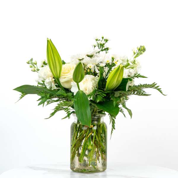 White lilies, roses, and other white flowers arranged in a clear glass jar vase