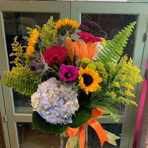Tall mixed arrangement of sunflowers, lilies, gerbera daisies, hydrangea in a glass vase with orange ribbon