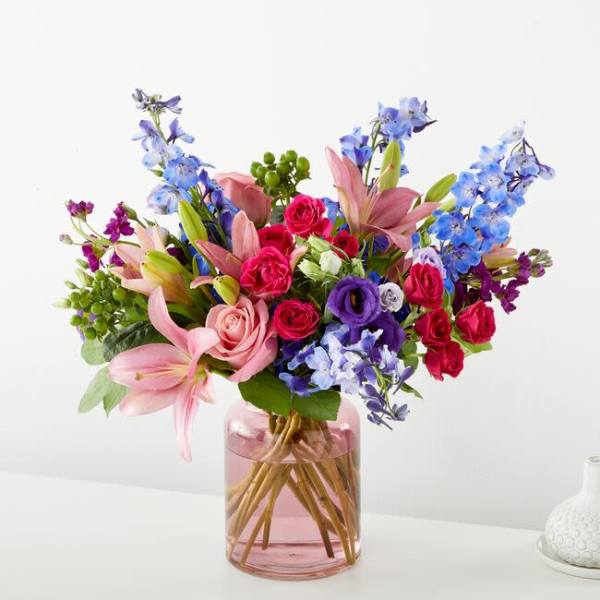 Mixed bouquet of pink roses, lilies, and blue flowers in a pink glass vase