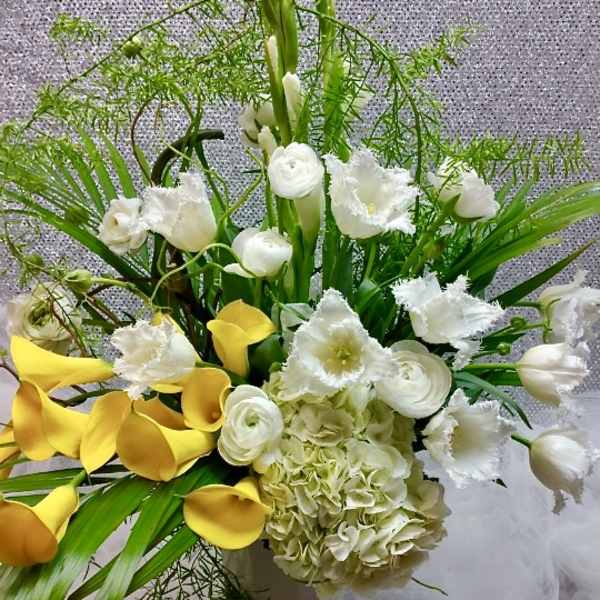 Tall arrangement of yellow calla lilies, white hydrangeas, tulips, and ranunculus in a white vase