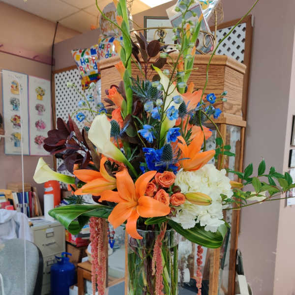 Tall mixed bouquet with orange lilies, blue accents, and white blooms in a glass vase