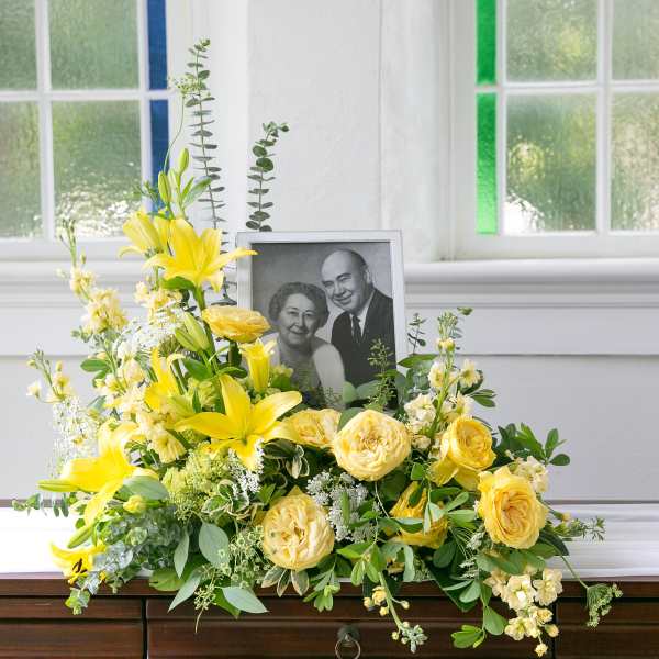 Yellow lily and rose sympathy arrangement displayed with a framed black-and-white portrait.