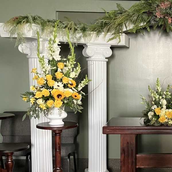 Yellow and white floral arrangements on pedestals beneath a white arch.