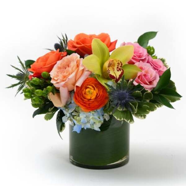 Low arrangement of orange and pink roses, green orchid, and mixed blooms in a clear glass cylinder vase.