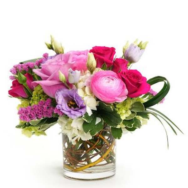 Low glass vase of pink roses, ranunculus, and mixed purple and white flowers in a compact arrangement