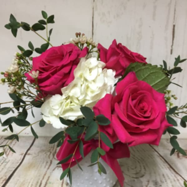 Small arrangement of hot pink roses and white hydrangea in a white textured vase with greenery.