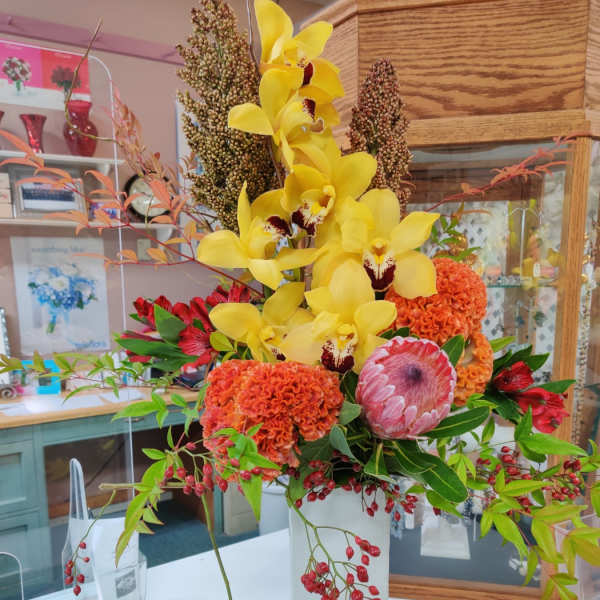 Tall arrangement with yellow orchids, orange blooms, pink protea and red berries in a white rectangular vase.