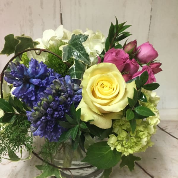 Short glass vase arrangement with yellow rose, pink roses, blue hyacinth, and white blooms