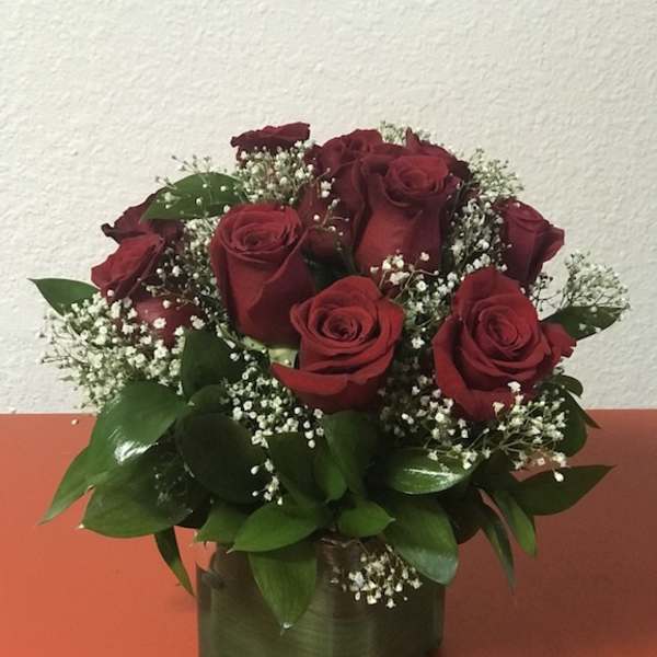 Compact arrangement of red roses with white filler flowers in a green cube vase on an orange surface