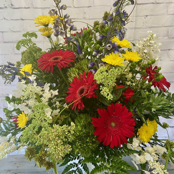 Mixed bouquet with red gerbera daisies, yellow blooms, and blue flowers in a white container