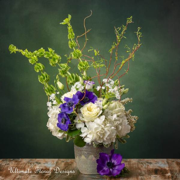 White and purple floral arrangement in a textured vase with tall curly branches.