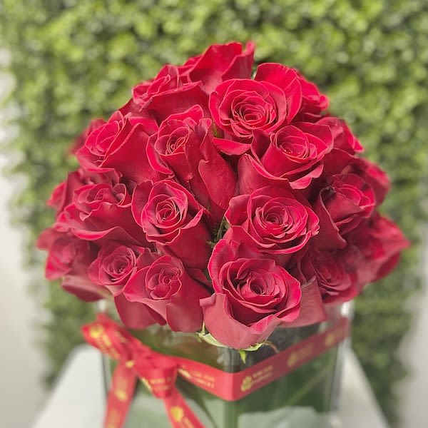 Dense bouquet of red roses arranged in a square glass vase wrapped with a red ribbon.
