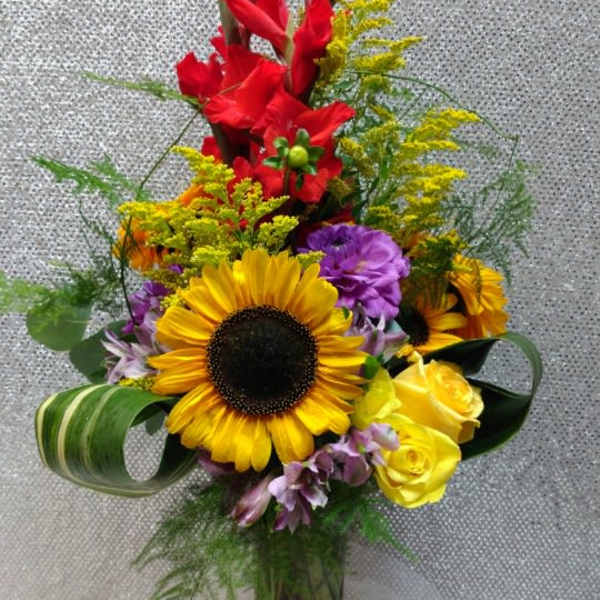 Tall mixed bouquet with sunflowers, red gladiolus, and yellow roses in a clear vase