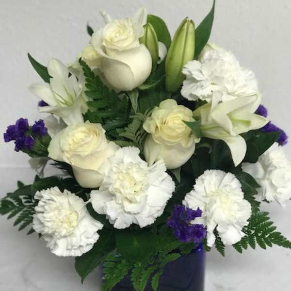 Compact arrangement of white roses, lilies, and carnations with purple accents in a dark blue cube vase