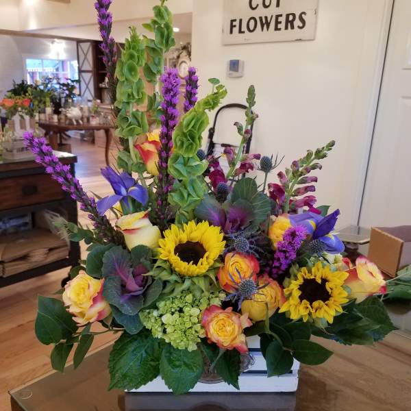 Bright mixed arrangement with sunflowers, roses, and purple flowers in a white wooden box.