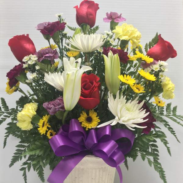 Mixed bouquet of roses, lilies, and daisies in a cream vase with a purple bow